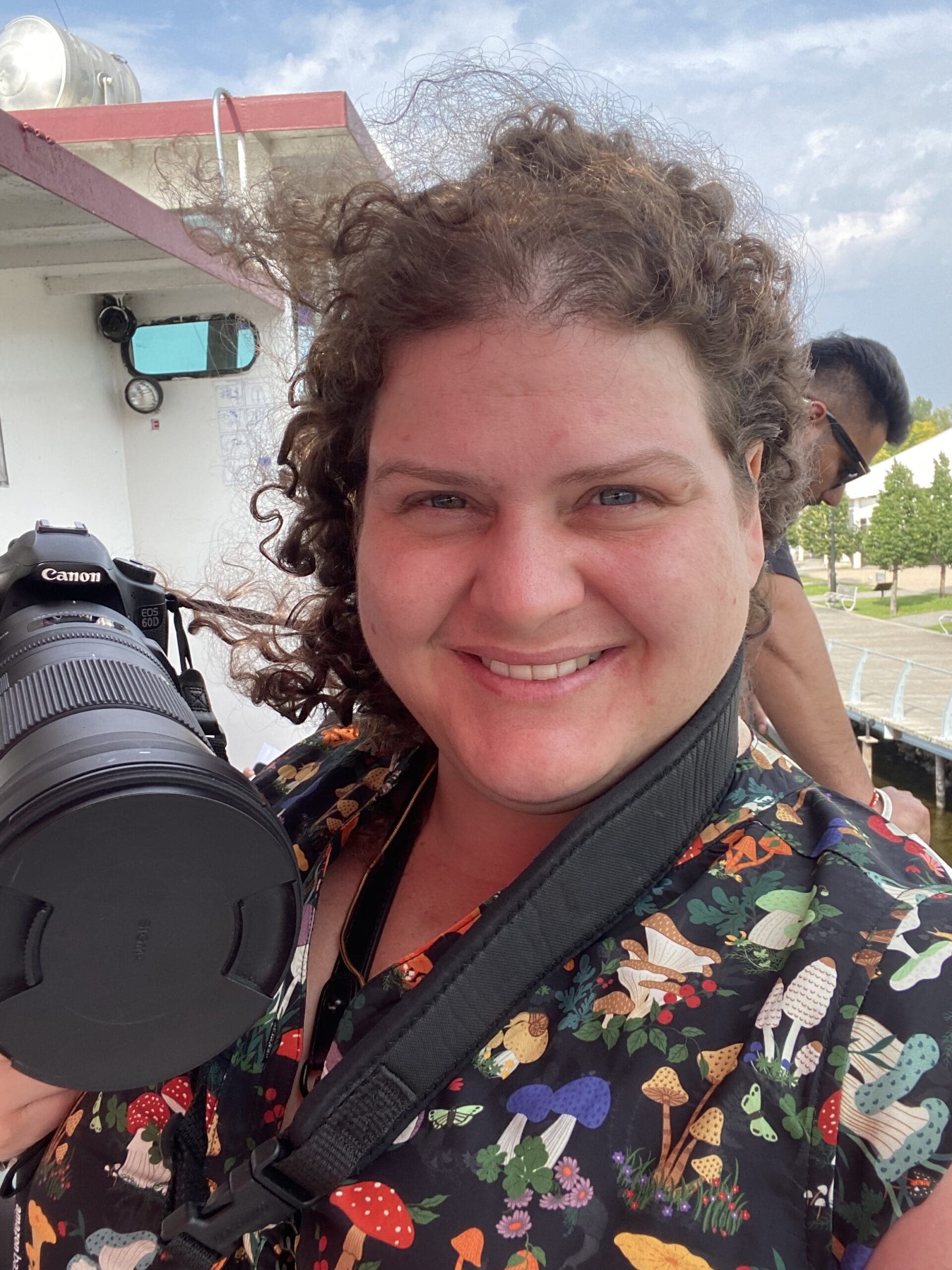 QuirkNSpite's creator Cassandra, the curls behind the camera, posing with her camera, lens on, in a quirky dress and a mischievous smile, she fully embraces the Quirky Lifestyle.